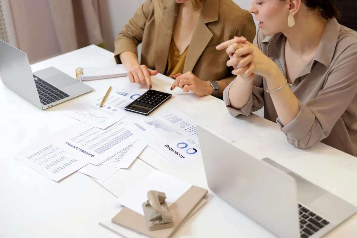 Two women at a desk reviewing accounting papers with a calculator and laptops