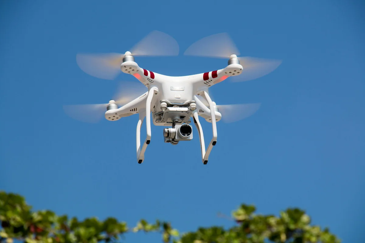 White camera drone hovering against a clear blue sky above green treetops